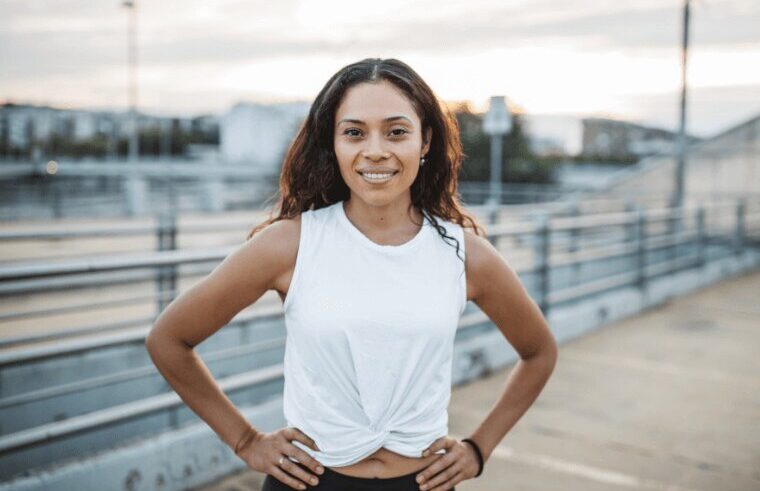 A woman with long curly hair, wearing a white sleeveless top and black leggings, stands outside with her hands on her hips, smiling at the camera. The background shows an urban setting with a railing and buildings.