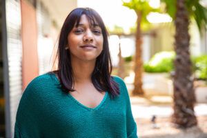 A woman with straight dark hair and bangs, wearing a teal sweater, stands outdoors and smiles slightly. Palm trees and sunlight are visible in the background, creating a warm, relaxed atmosphere.