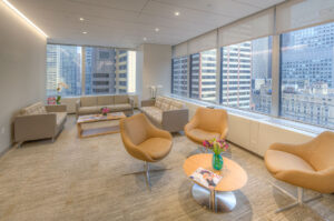 Modern office lounge with tan armchairs, sofas, coffee tables with magazines and flowers, large windows overlooking city skyscrapers, and neutral-toned décor.