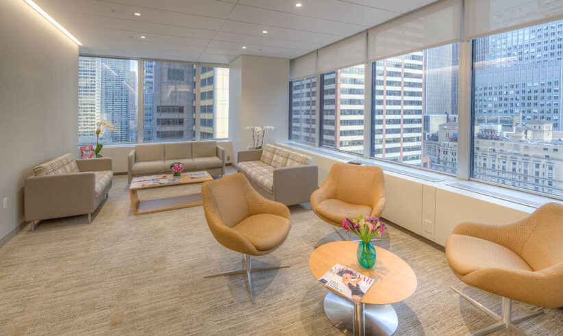 Modern office lounge with tan armchairs, sofas, coffee tables with magazines and flowers, large windows overlooking city skyscrapers, and neutral-toned décor.