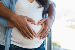 Close-up of a pregnant woman’s belly with two pairs of hands gently forming a heart shape over it, symbolizing love, support, and anticipation for the baby.