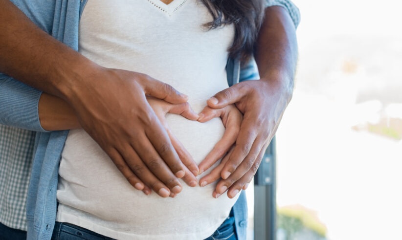 Close-up of a pregnant woman’s belly with two pairs of hands gently forming a heart shape over it, symbolizing love, support, and anticipation for the baby.