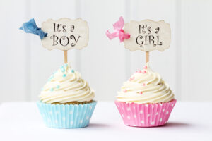 Two cupcakes with white frosting: one in a blue polka dot wrapper with a sign reading Its a Boy, and one in a pink polka dot wrapper with a sign reading Its a Girl. Both have matching colored bows and sprinkles.