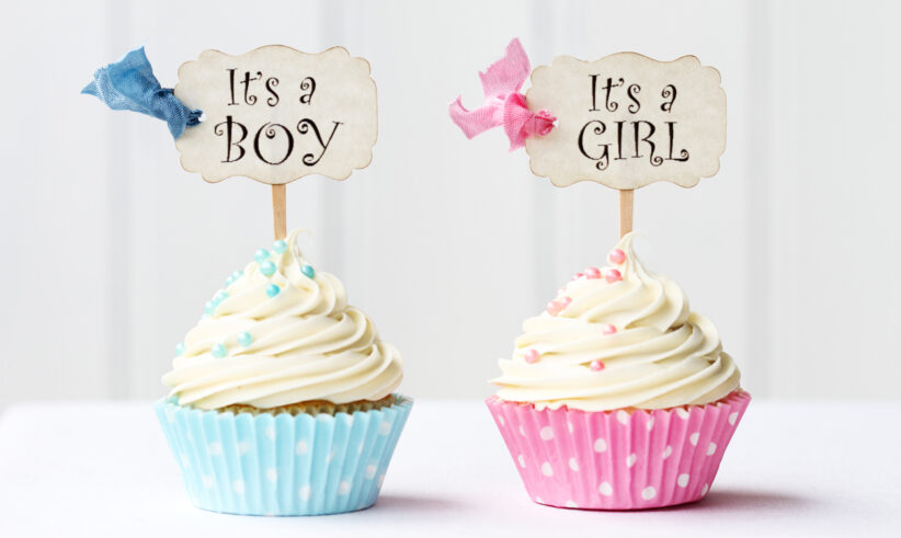 Two cupcakes with white frosting: one in a blue polka dot wrapper with a sign reading Its a Boy, and one in a pink polka dot wrapper with a sign reading Its a Girl. Both have matching colored bows and sprinkles.