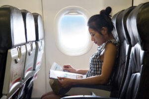 A woman sits in an airplane seat by the window, reading a magazine. Sunlight streams through the window beside her, and the cabin appears calm and quiet.