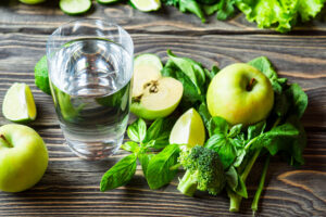 A glass of water sits on a wooden table surrounded by green apples, spinach leaves, broccoli, lime wedges, basil, and leafy greens. The fresh produce creates a healthy and vibrant arrangement.