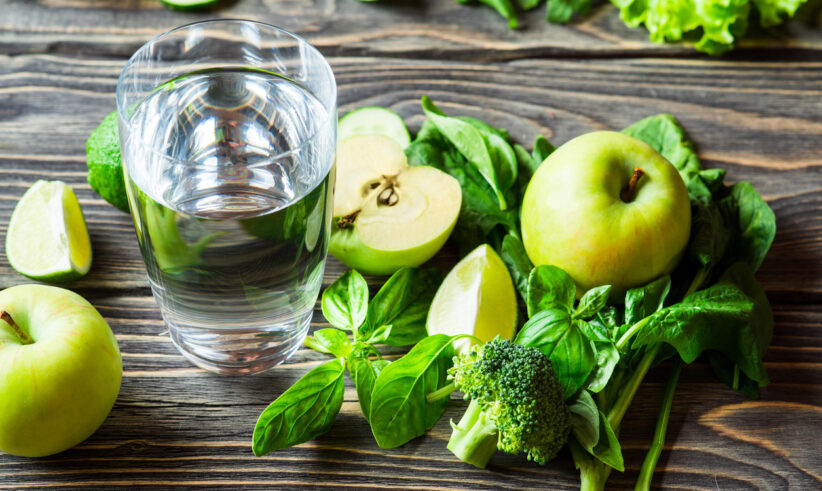 A glass of water sits on a wooden table surrounded by green apples, spinach leaves, broccoli, lime wedges, basil, and leafy greens. The fresh produce creates a healthy and vibrant arrangement.