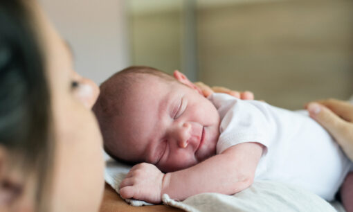 A newborn baby wearing a white onesie sleeps peacefully on an adult’s chest, with the adult’s face partially visible in the foreground. The background is softly blurred.