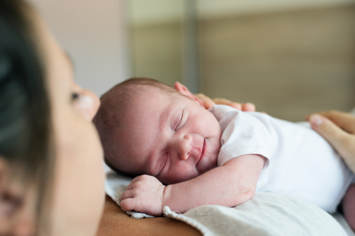 A newborn baby wearing a white onesie sleeps peacefully on an adult’s chest, with the adult’s face partially visible in the foreground. The background is softly blurred.