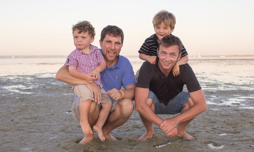 Two men and two young boys pose and smile together on a sandy beach during sunset. The men crouch down, with one boy sitting on each of their knees. The background shows calm water and a clear sky.