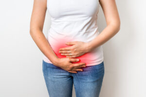 A person wearing a white shirt and blue jeans holds their lower abdomen with both hands; a red highlight on the area suggests pain or discomfort.