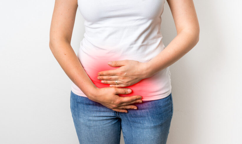 A person wearing a white shirt and blue jeans holds their lower abdomen with both hands; a red highlight on the area suggests pain or discomfort.