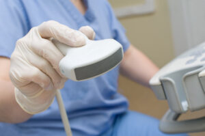 A close-up of a healthcare worker wearing gloves and holding an ultrasound probe, with part of the ultrasound machine visible in the background.