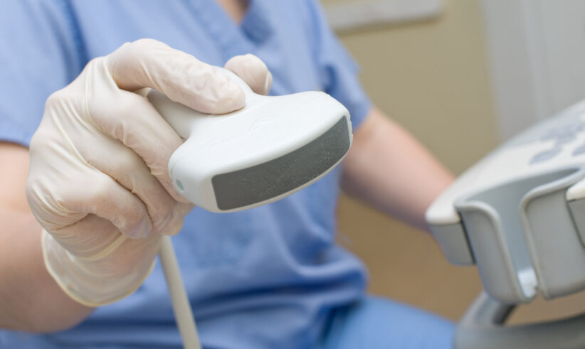 A close-up of a healthcare worker wearing gloves and holding an ultrasound probe, with part of the ultrasound machine visible in the background.