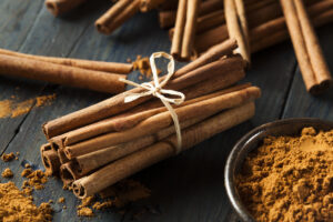 A bundle of cinnamon sticks tied with twine sits on a wooden surface next to a bowl of ground cinnamon, with more cinnamon sticks and scattered powder in the background.
