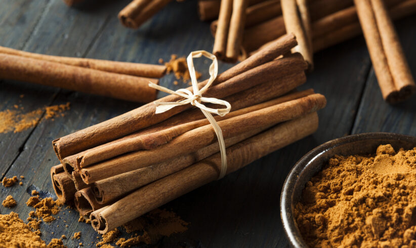 A bundle of cinnamon sticks tied with twine sits on a wooden surface next to a bowl of ground cinnamon, with more cinnamon sticks and scattered powder in the background.