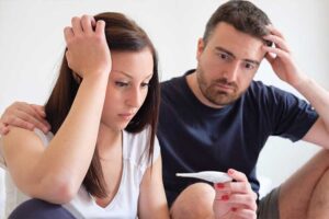 A worried-looking couple sits close together, both touching their heads with concerned expressions, as the woman holds and looks at a pregnancy test.