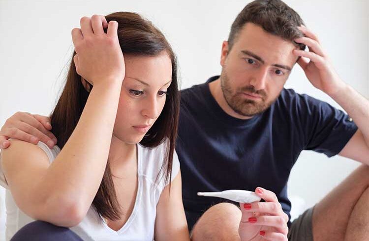 A worried-looking couple sits close together, both touching their heads with concerned expressions, as the woman holds and looks at a pregnancy test.