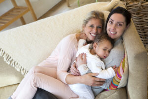 Two women sit closely together on a beige couch, smiling at the camera, while holding a sleeping baby in their arms. The scene appears warm and affectionate, with soft natural light.
