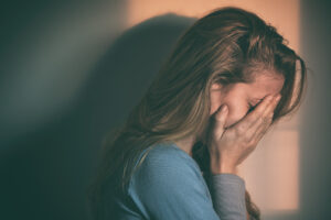 A woman with long hair covers her face with her hand, appearing upset or distressed, while standing in dim lighting with a shadow cast on the wall behind her.