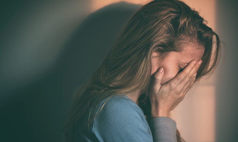 A woman with long hair covers her face with her hand, appearing upset or distressed, while standing in dim lighting with a shadow cast on the wall behind her.
