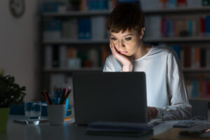 A woman sits at a desk late at night, looking intently at her laptop screen with her hand on her face. The room is dimly lit, and there are office supplies and a glass of water on the desk.