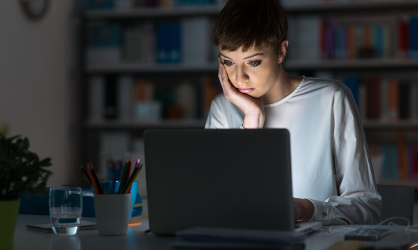 A woman sits at a desk late at night, looking intently at her laptop screen with her hand on her face. The room is dimly lit, and there are office supplies and a glass of water on the desk.