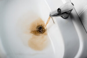 Brown, dirty water flows from a faucet into a white sink, swirling around the drain, indicating contaminated or rusty tap water.