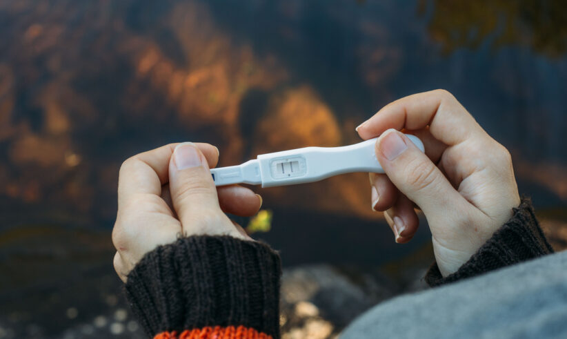 Close-up of hands holding a pregnancy test with one line visible, indicating a negative result. The person is wearing a dark sweater with an orange sleeve, and there is a blurred outdoor background.