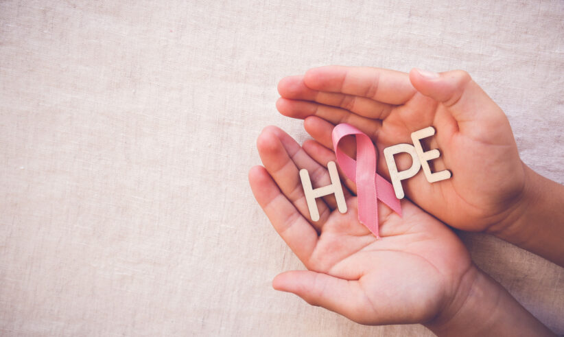 Two hands gently hold wooden letters spelling “HOPE” with a pink ribbon, symbolizing breast cancer awareness, on a light fabric background.