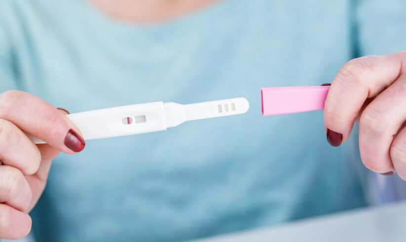 A person holding a pregnancy test with two visible lines, indicating a positive result. The person’s fingernails are painted red and they are wearing a light blue shirt.
