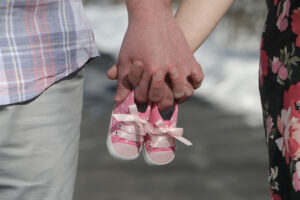 Two people holding hands are holding a pair of small pink baby shoes. One person is wearing light pants and a plaid shirt; the other is wearing a floral dress. The background is blurred.