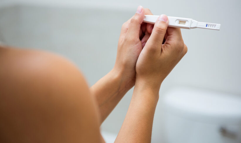 A person is holding a pregnancy test with both hands, looking at the result. The background is out of focus, showing a bathroom setting.