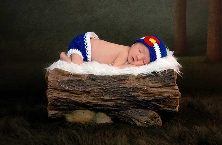 A newborn baby sleeps on a white fuzzy blanket atop a log prop, wearing a blue knit hat with a red and yellow accent and matching blue diaper cover, set against a dark forest-themed background.