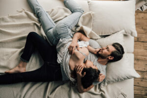 A couple lies on a bed, cuddling closely. The man is on his back, gently holding the woman, who rests her head on his chest. They appear relaxed and comfortable, surrounded by pillows and a light blanket.