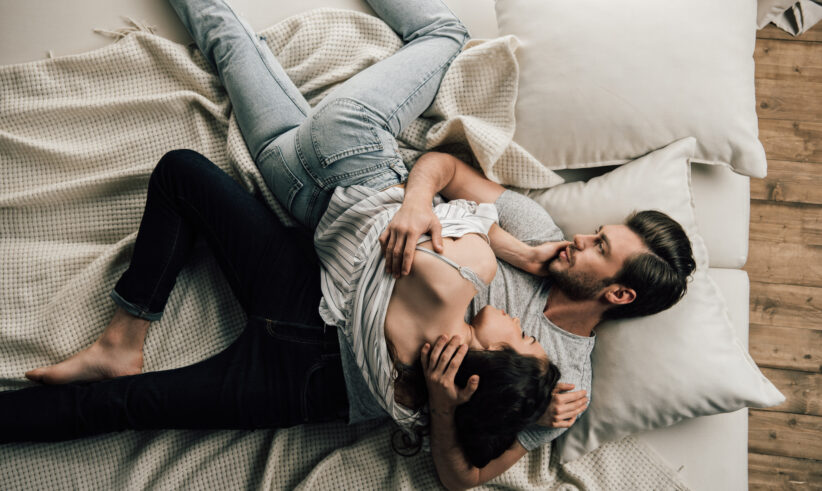 A couple lies on a bed, cuddling closely. The man is on his back, gently holding the woman, who rests her head on his chest. They appear relaxed and comfortable, surrounded by pillows and a light blanket.