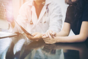 A doctor in a white coat discusses medical results with a patient, holding a paper while the patient clasps their hands together on a table, suggesting a comforting or supportive conversation.