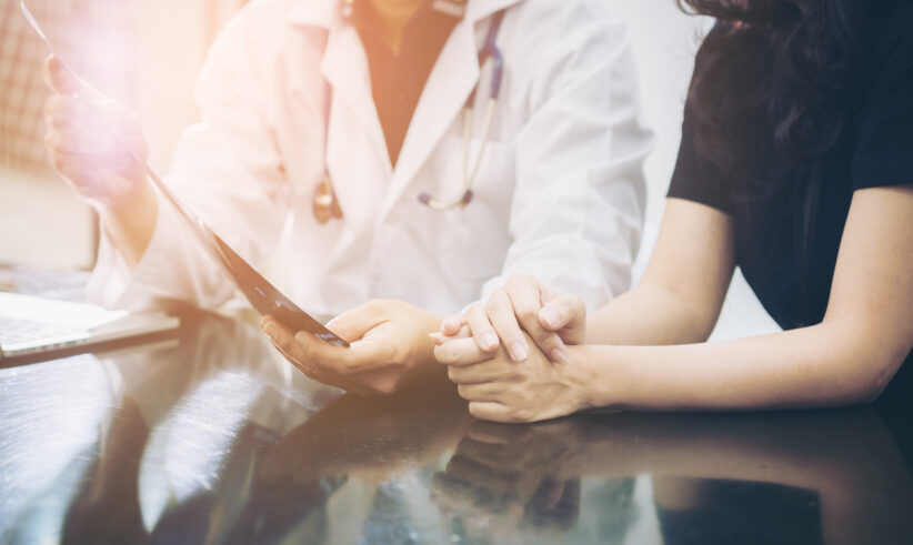 A doctor in a white coat discusses medical results with a patient, holding a paper while the patient clasps their hands together on a table, suggesting a comforting or supportive conversation.