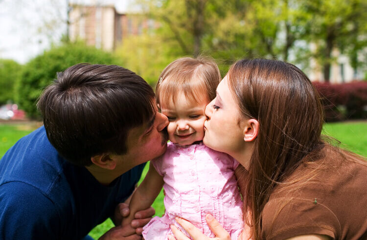 A mother and father kiss their smiling baby daughter on the cheeks while sitting on grass in a park, with trees and a building in the background.