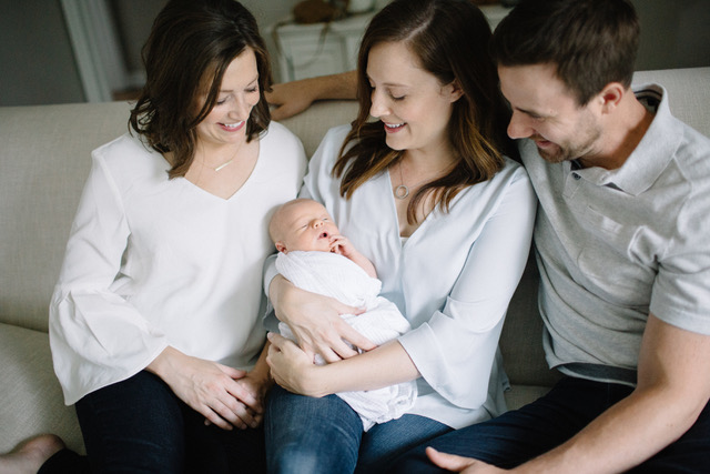 Three adults sit close together on a couch, smiling and looking at a newborn baby wrapped in a white blanket, held by the woman in the center. The atmosphere is warm and joyful.