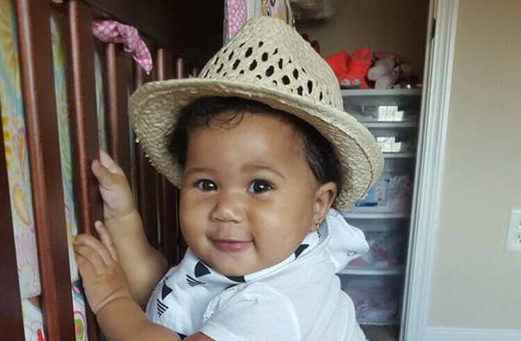 A smiling baby wearing a straw hat and a white outfit stands holding onto a crib rail in a cozy, well-lit room.