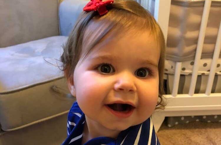 A smiling baby with brown hair, wearing a blue and white striped shirt and a red bow hair clip, sits in front of a white crib and a beige chair.
