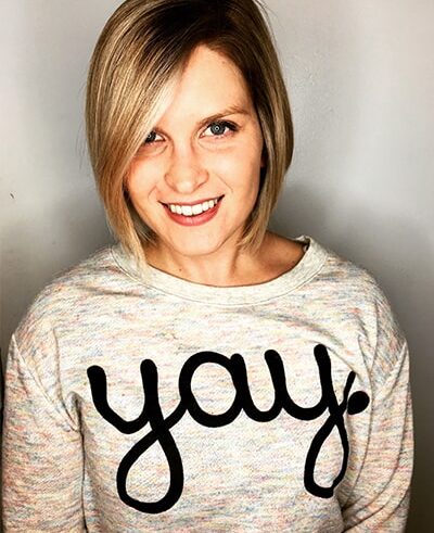 A smiling woman with straight blonde hair wears a light-colored sweater with the word yay. written in large black script across the front, standing against a plain background.