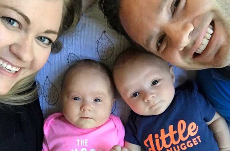 A smiling woman and man lie next to two babies. The baby on the left wears a pink onesie, and the baby on the right wears a blue onesie that says Little Nugget. They are all lying close together, looking at the camera.
