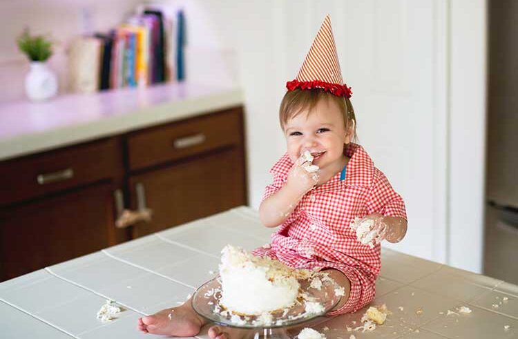 A smiling toddler in a red checkered outfit and party hat sits on a kitchen counter, happily eating and playing with a smashed birthday cake, with cake smeared on their face and hands.