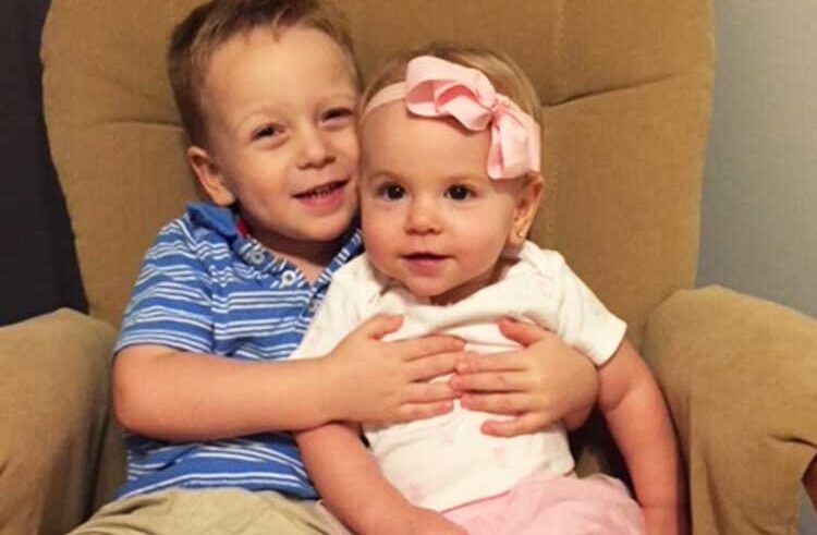 A young boy in a blue striped shirt smiles and hugs a baby girl wearing a pink headband and white shirt as they sit together on a tan armchair.