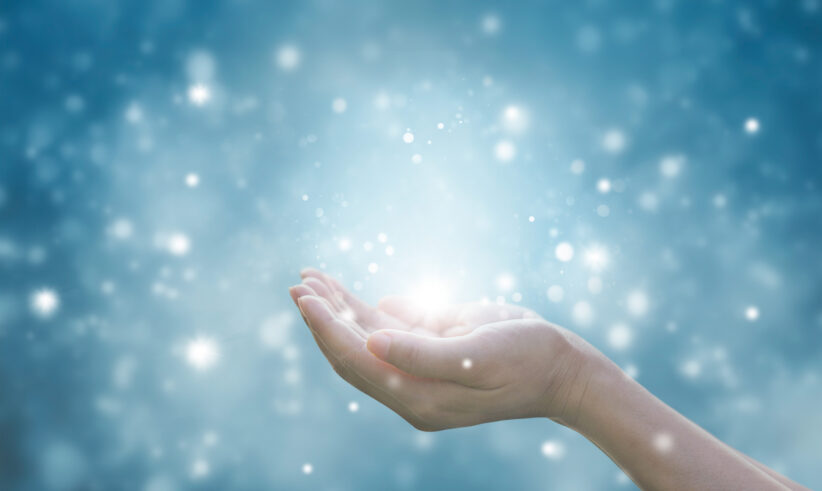 A hand gently cupped and extended against a blue background, surrounded by glowing, ethereal light and sparkling particles, creating a magical or hopeful atmosphere.