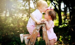 Two young children in pink clothes stand on a wooden ladder outdoors. One child kisses the other on the forehead, both smiling, with green trees and sunlight in the background.