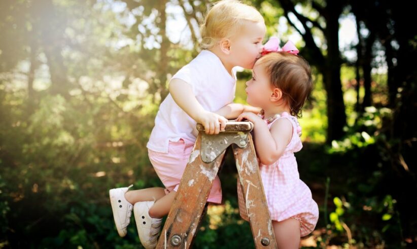 Two young children in pink clothes stand on a wooden ladder outdoors. One child kisses the other on the forehead, both smiling, with green trees and sunlight in the background.