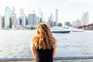 A person with curly hair, seen from behind, stands by a railing overlooking a river with a city skyline and a boat in the background on a sunny day.